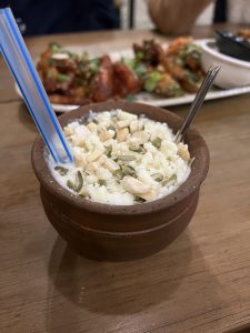 A clay pot filled with a white yogurt drink is topped with nuts and seeds, served with a blue-striped straw.