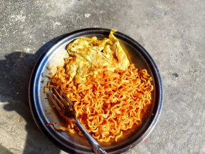 
A stainless steel plate with a serving of spicy instant noodles next to a partially cooked egg. 