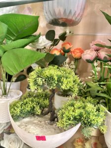 A display of artificial plants, featuring a small, decorative tree with vibrant green foliage set in a round white pot filled with a sandy texture. 