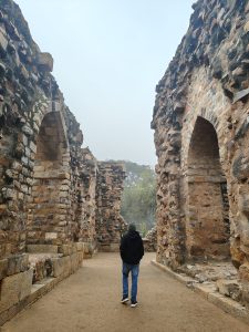 
A person wearing a black hoodie and jeans walks along a pathway between stone ruins, characterized by weathered brick walls and arches