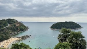 A stunning coastal overlook showing a sandy beach cove on the left and a small, tree-covered island on the right, surrounded by clear turquoise water.