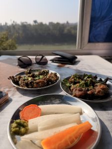 Two metal plates are set on a table, one with assorted vegetable pieces including carrots and white radish, accompanied by green chutney. The other plate contains cooked meat pieces garnished with green herbs.