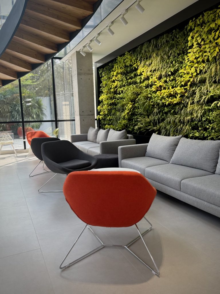 An interior wall on the right covered in greenery. In front of the wall are two couches. Facing the couches are four chairs that look like scoops. A curving staircase is above, and a glass wall on the far end of the room.