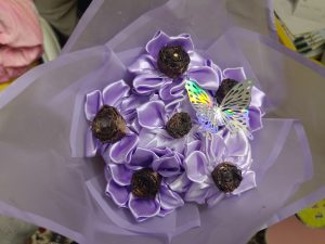 A bouquet featuring large, lavender satin ribbon flowers arranged amidst small, dark brown decorative items resembling flower buds.
