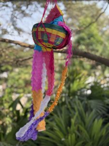 A colorful woven basket with vibrant streamers hanging from it, suspended outdoors with foliage in the background.
