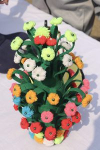 A vibrant decorative arrangement featuring a green base designed to resemble a vase, adorned with colorful felt flowers in various shades, including red, yellow, white, pink, and orange.