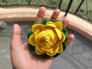 A person’s hand is holding a decorative flower made of yellow petals arranged in a circular pattern on a green base