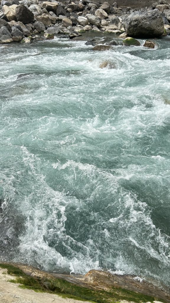 A turbulent river flows swiftly, its clear turquoise water creating frothy white caps as it moves over rocks and boulders.