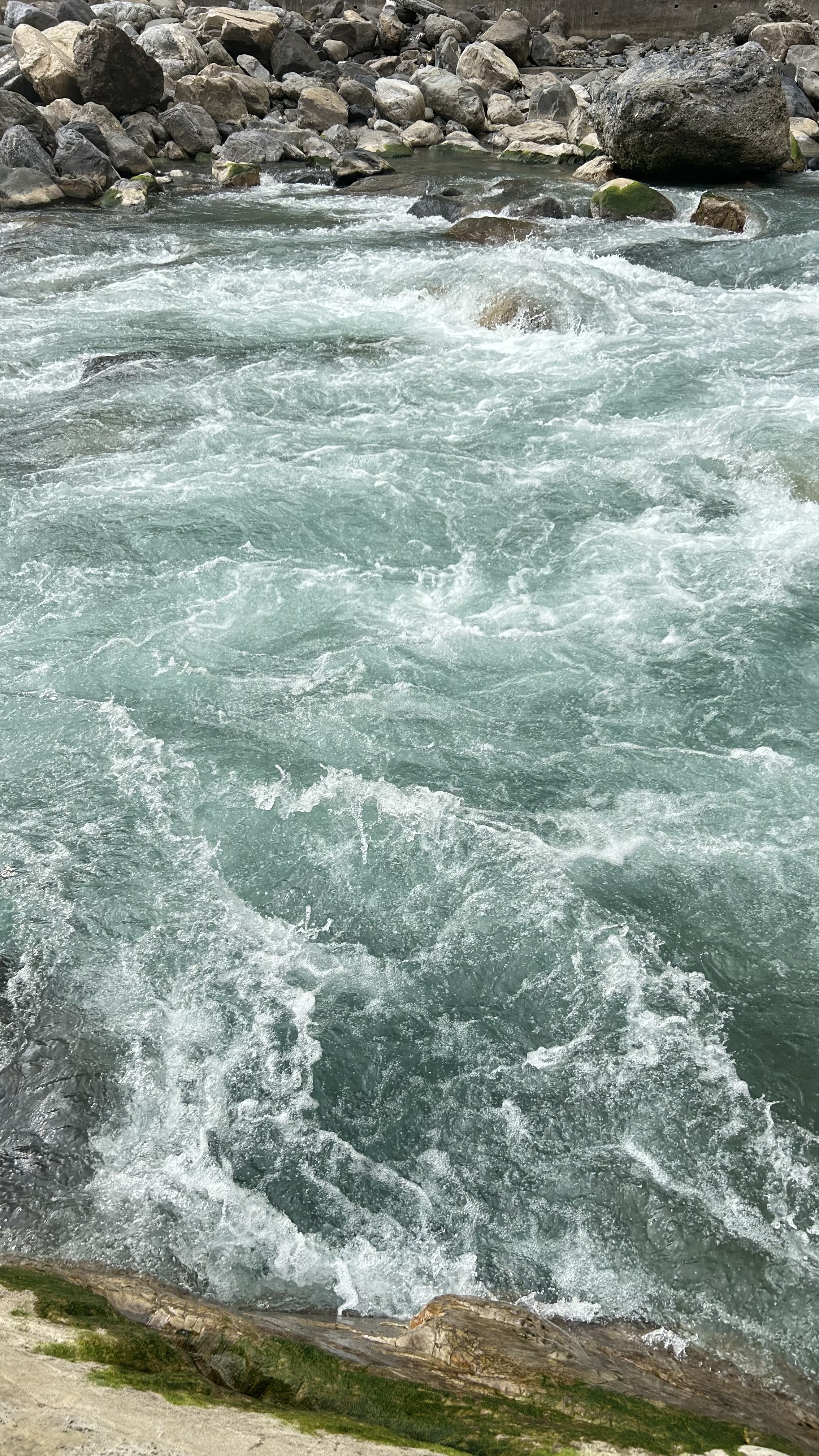 A turbulent river flows swiftly, its clear turquoise water creating frothy white caps as it moves over rocks and boulders.
