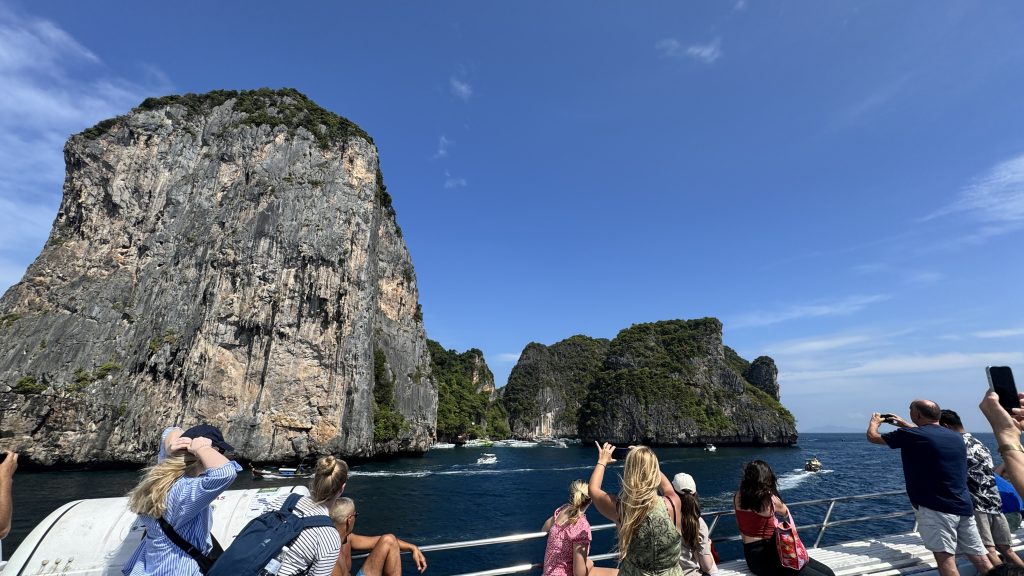 Tourists on the deck of a ferry taking photos of towering limestone cliffs and surrounding boats in bright blue water.