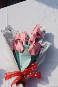 A bouquet of handmade paper flowers featuring pink rose-shaped blooms with light blue leaves. The bouquet is wrapped in a decorative layer that includes newspaper clippings, and it is tied with a red ribbon adorned with white hearts