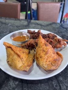 A white plate is served on a dark marble surface, featuring two golden-brown samosas with a crispy texture on the left and a portion of deep-fried chicken pieces on the right.