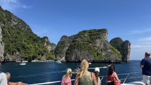 Passengers on the deck of a boat admiring a beautiful cove surrounded by massive, lush green limestone cliffs and clear turquoise water.