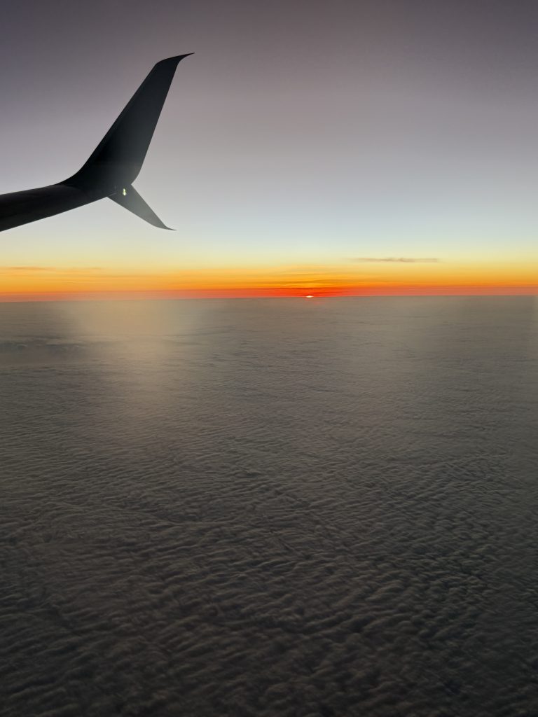 A sunset view from an airplane window, with the silhouette of the airplane’s wing in the foreground.