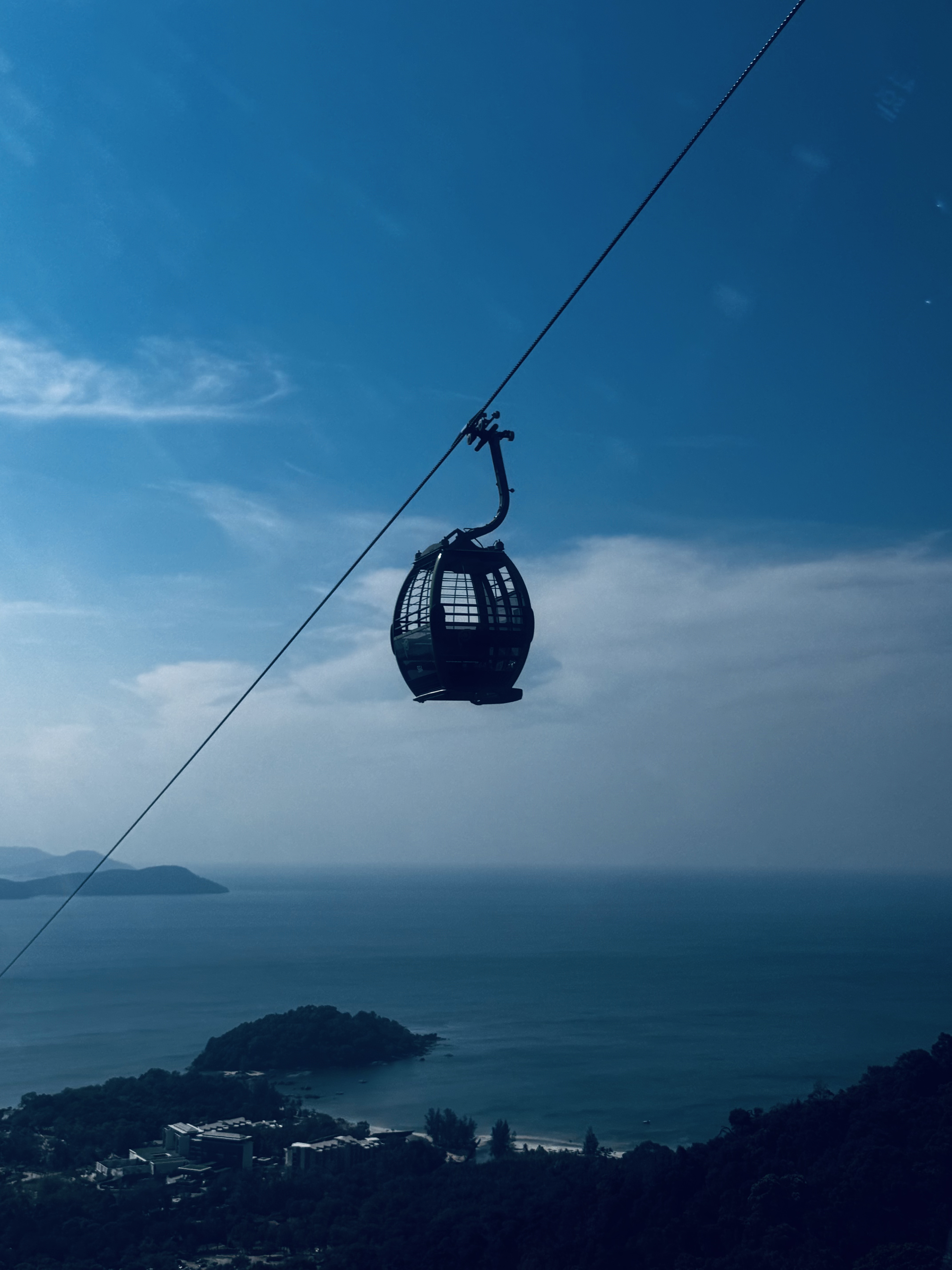 A single gondola is suspended high above a vast body of water, traveling along a cable. The background features gentle rolling hills and a distant island with lush greenery. The sky above is cloudy.