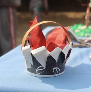 A handmade paper crown displayed on a blue tablecloth, featuring a black base with white accents and red fabric strips emerging from the top. 