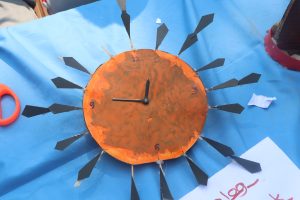 A handmade clock with an orange circular face and black hour markers positioned around its edge. 