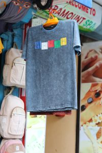 A t-shirt hangs on an orange hanger at a street shop, featuring a design of five colorful nepali prayer flags