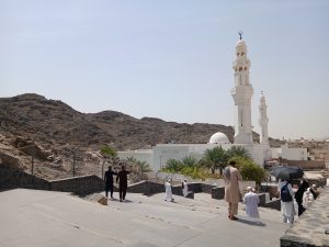 A daytime view of a mosque located near rocky hills, with visitors walking down stone steps toward the prayer area. Palm trees, white minarets, and surrounding mountains create a calm and spiritual atmosphere. The image captures pilgrims and visitors in a natural and architectural landscape.