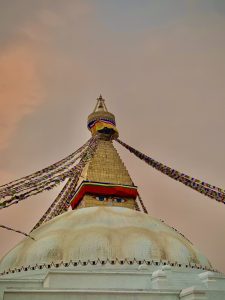 The top of a stupa with colorful prayer flags, a golden spire, and painted eyes, set against a soft pink sky.