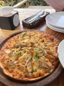 A wooden serving board holds a freshly baked pizza topped with melted cheese, sliced grilled chicken, and fresh arugula. 