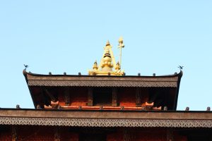 An intricate traditional roof with wooden carvings and a golden stupa-like pinnacle, with a small bird perched on the edge against a clear blue sky.