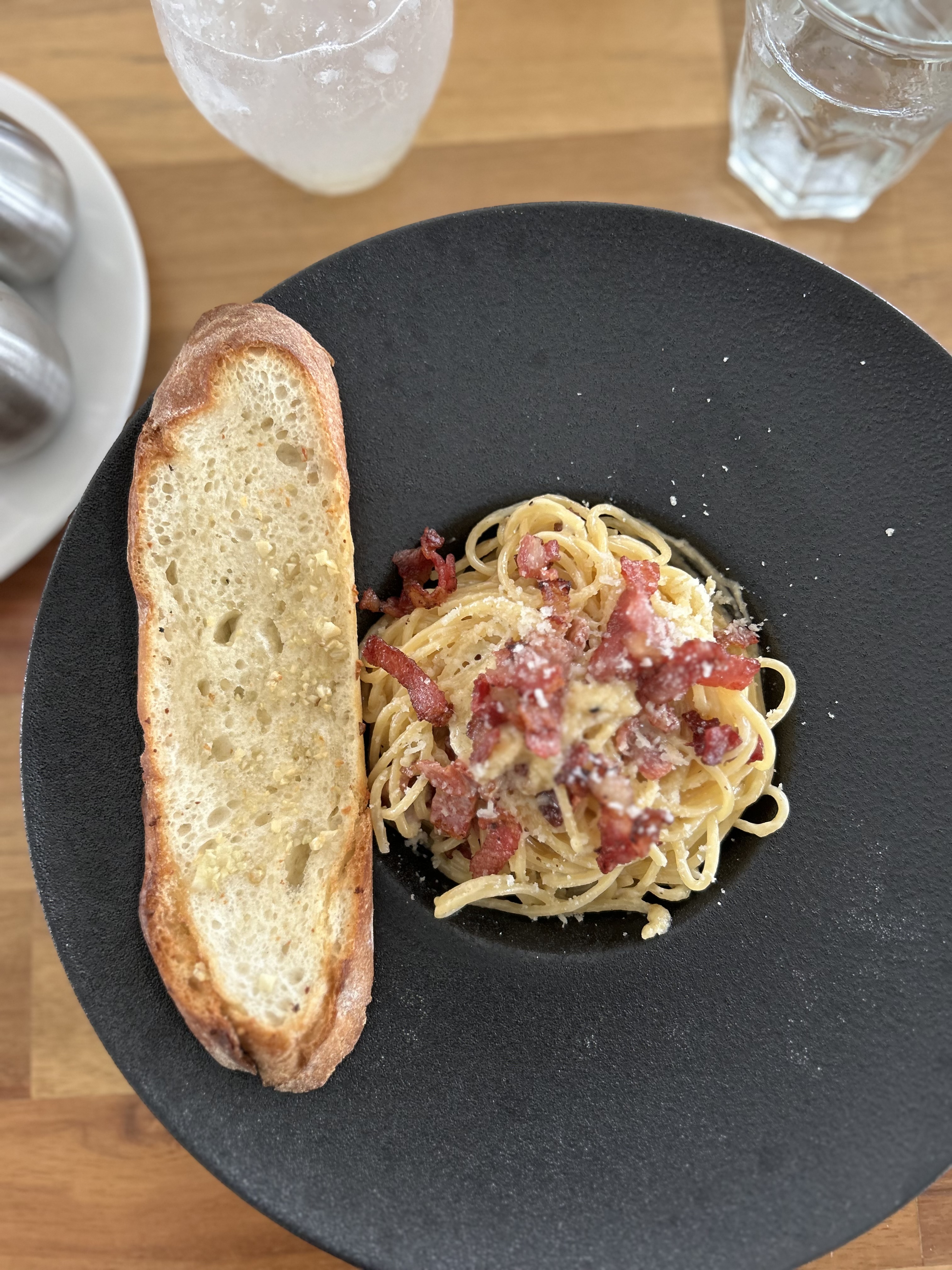 A black, round plate features a serving of spaghetti topped with small pieces of crispy bacon and sprinkled with grated cheese.