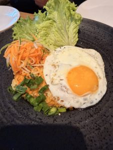 
A black plate holds a serving of fried rice topped with a sunny-side-up egg, featuring a bright yellow yolk. Beside the rice, there is a serving of pickled shredded carrots and onions, garnished with chopped green onions. Curved lettuce leaves provide a fresh contrast to the dish.