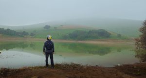 
A person wearing a helmet stands on the edge of a calm lake, gazing at the reflective water surrounded by lush green hills shrouded in mist. 
