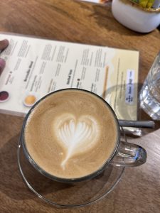 Closeup of the top of a cup of coffee.  There's foam on top with the shape of a heart on it. In the background, out of focus, are a menu, glass, and other table top things.