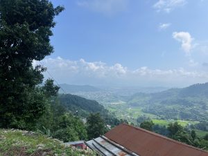 A scenic view from a hillside, featuring lush green valleys and distant mountains under a partly cloudy blue sky.