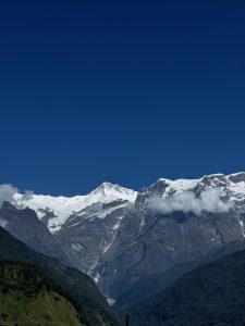 
A panoramic view of snow-capped mountains under a clear blue sky, featuring jagged peaks, some partially obscured by wispy clouds, and lush greenery in the foreground. T