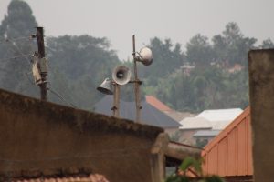 A close-up view of two loudspeakers mounted on wooden poles, with rooftops and dense trees in the background on a cloudy day – traditional radio in many remote areas of Uganda
