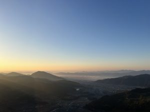 A panoramic view of a landscape at sunrise, showcasing rolling hills and distant mountains under a clear blue sky. 