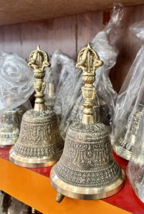 
A collection of intricately designed brass bells displayed on a wooden shelf. Each bell features ornate patterns and a decorative handle at the top, partially covered in plastic wrap. 