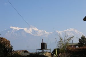 A panoramic view of snow-capped mountains under a clear blue sky, with a small water tank and foliage in the foreground
