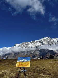 A vibrant painting of a snow-capped mountain range is displayed on an easel in a grassy meadow. The background features towering peaks partially shrouded in clouds under a bright blue sky