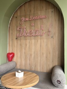 A cozy seating area with a wooden wall, a pink neon “You Deserve a Treat!” sign, a round table, and a cushioned bench.