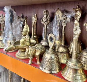 
A display of various brass artifacts on a shelf, including decorative bells and animal figures. Prominently featured are a small brass cow and elephant, alongside intricately designed bells adorned with different motifs.
