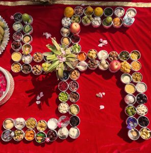 Dozens of small silver bowls filled with various fruits, nuts, and grains are arranged in a swastika shape on a red cloth for weaning ceremony of a baby.
