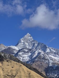 
A snow-capped mountain fishtail peak rises sharply against a clear blue sky, with a few fluffy white clouds scattered across the scene.