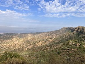 Panoramic view of rugged hills with sparse greenery, overlooking the coastline and calm ocean under a partly cloudy sky.