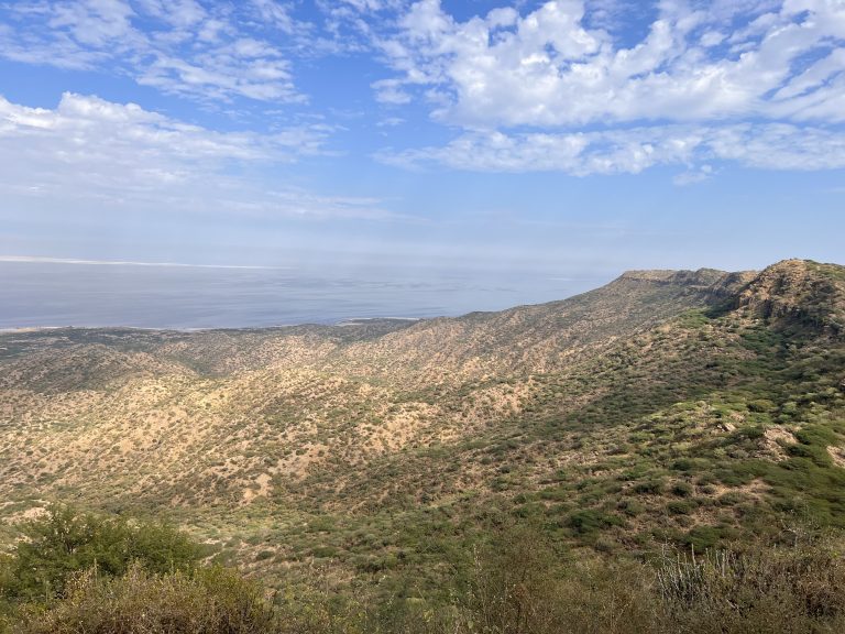 Panoramic view of rugged hills with sparse greenery, overlooking the coastline and calm ocean under a partly cloudy sky.