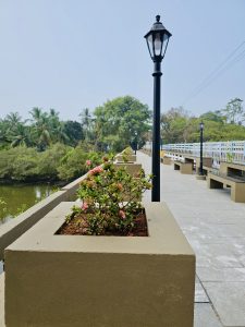 A peaceful walkway with lamps, benches, and plants beside a quiet river in Kallampara, Feroke, Kozhikode. The greenery and open sky create a relaxing mood.
