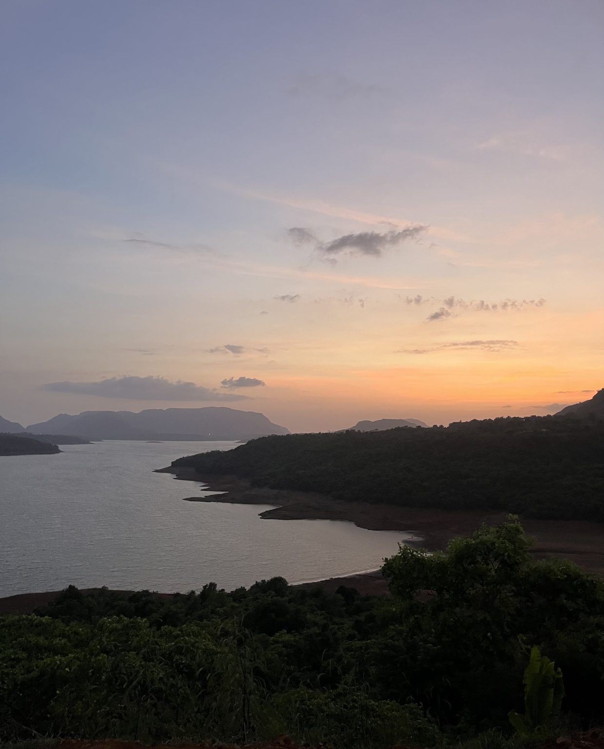 A wide, high-angle landscape shot of a calm lake winding through rolling green hills at dusk. The sky transitions from a soft blue at the top to a warm orange and yellow glow on the horizon where the sun has set. Dark, silhouettes of forested land and distant mountains frame the water, creating a serene and quiet atmosphere.