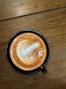 A top-down view shows a cup of latte with leaf-patterned latte art and a dusting of cinnamon on a wooden surface
