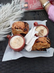 A wooden serving tray holds two small circular cups filled with a frothy beverage, possibly coffee, alongside a piece of toasted bread with a charred crust