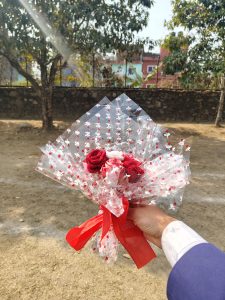 A bouquet of red fabric roses wrapped in clear cellophane with a red ribbon, set against trees and buildings.
