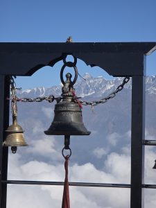 A close-up view of two bells hanging from a metal structure, with a picturesque backdrop of snow-capped mountains under a clear blue sky.