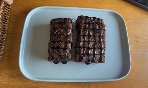 A light blue plate holds two thick, rectangular brownies cut into squares, drizzled with a glossy chocolate sauce.